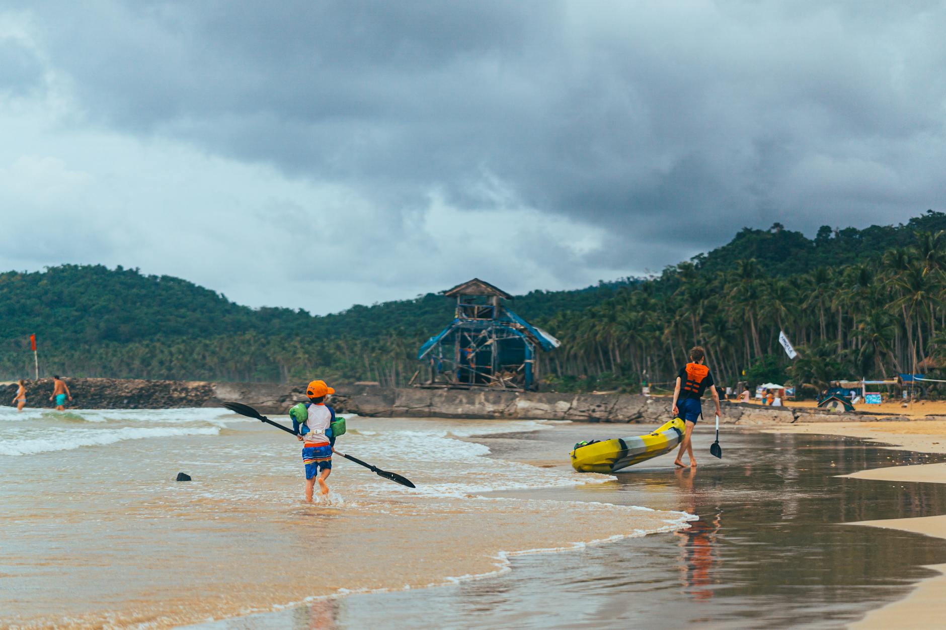 Dwoje dzieci z kajakami na piaszczystej plaży pod zachmurzonym niebem